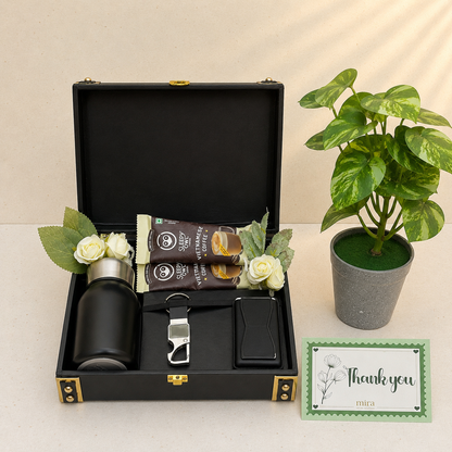 A black gift box set opened to reveal a black bottle, two packaged items, a silver opener, a black card holder, and white flowers. A potted green plant and a small thank you card sit beside the box on a light beige surface.