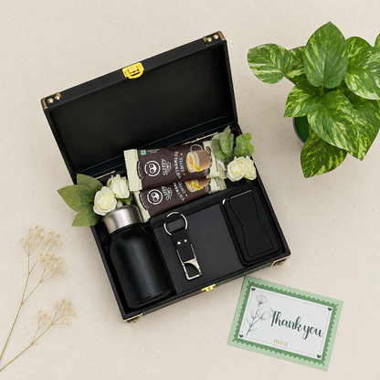 A black presentation gift box opened on a light beige surface, containing a black bottle, two brown packaged items, a metal bottle opener, a black card holder, and small white flowers. A green potted plant and a thank you card are placed beside the box.