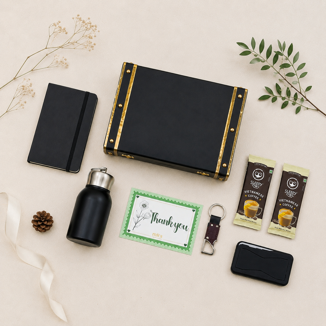 A curated gift set arranged on a light beige background with decorative dried flowers and greenery. The set includes a black hard case with gold trim, a black notebook, a black insulated bottle with a silver cap, a green thank-you card, two packaged coffee packets, a small black card holder, a keychain, and a pinecone.