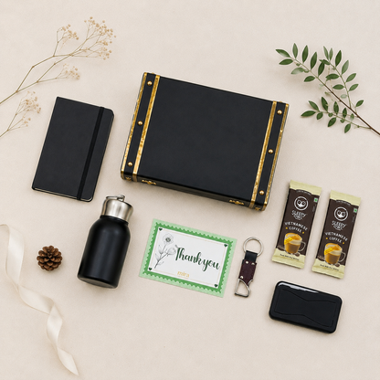 A curated gift set arranged on a light beige background with decorative dried flowers and greenery. The set includes a black hard case with gold trim, a black notebook, a black insulated bottle with a silver cap, a green thank-you card, two packaged coffee packets, a small black card holder, a keychain, and a pinecone.