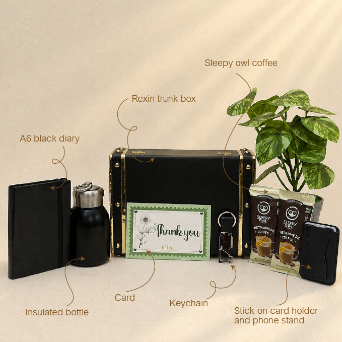 A curated gift set arranged on a beige background with handwritten callouts naming each item: A6 black diary, insulated bottle, Rexin trunk box, card, keychain, MagSafe card holder and phone stand, and Sleepy owl coffee. The main product is the black trunk box with gold-tone hardware, surrounded by matching accessories.
