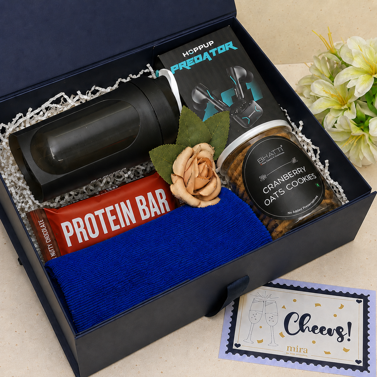A curated gift box set in a black presentation box with white crinkle paper filler, containing a black insulated bottle, a red protein bar, a blue cloth, a jar of cranberry oats cookies, a black card holder, and a small peach rose with green leaves. A small card reading 'Cheers! mita' is placed outside the box on a light beige surface with white flowers in the background.